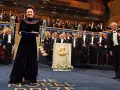 Uroczystość wręczenia Nagrody Nobla. W centrum kobieta w granatowej sukni z dyplomem. W tle orkiestra i laureaci. Tokarczuk nobel za co? Za literaturę.