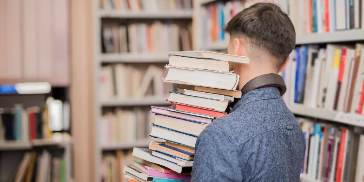Mężczyzna w bibliotece niesie stos książek, jakby szukał streszczenia "Nad Niemnem".