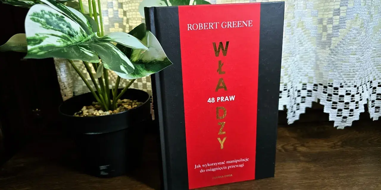 Książka "48 praw władzy" Roberta Greene'a na drewnianej półce, obok rośliny w czarnej doniczce.