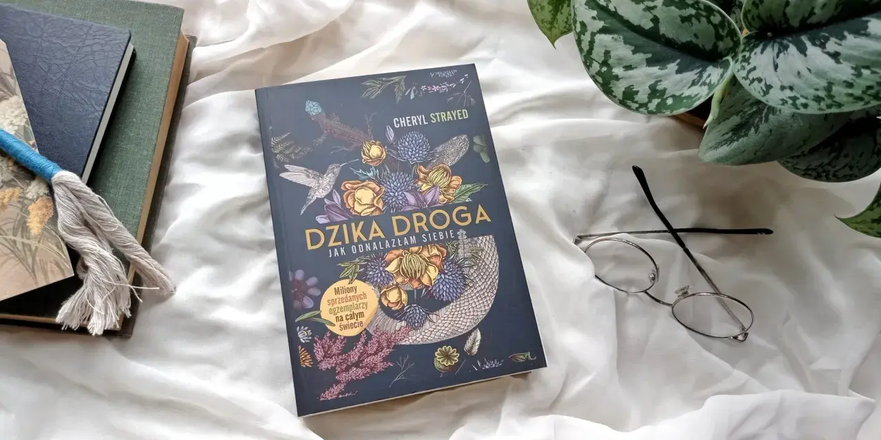 Okładka książki "Dzika droga" z motywami roślinnymi i zwierzęcymi. Obok leżą okulary i inne książki.