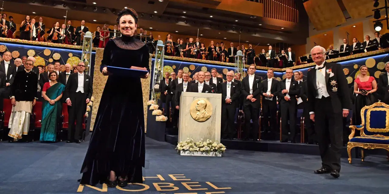 Uroczystość wręczenia Nagrody Nobla. W centrum kobieta w granatowej sukni z dyplomem. W tle orkiestra i laureaci. Tokarczuk nobel za co? Za literaturę.