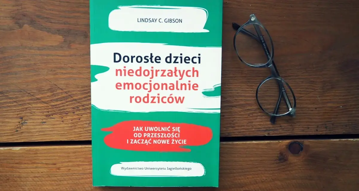 Książka "Dorosłe dzieci niedojrzałych emocjonalnie rodziców" i okulary na drewnianym tle.