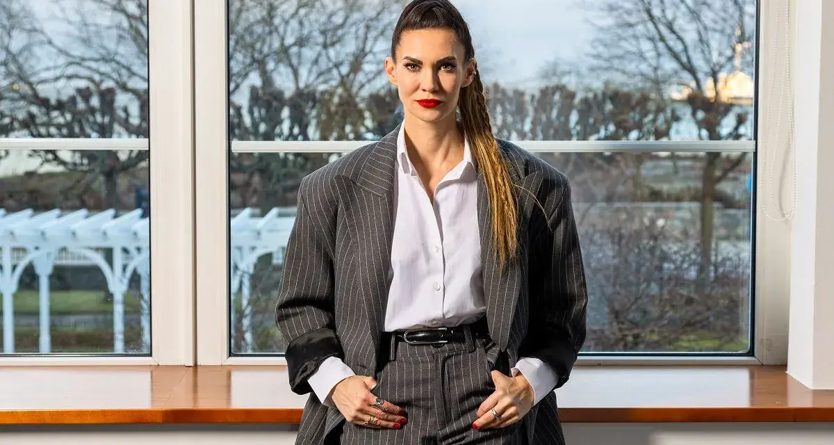 Małgorzata Oliwia Sobczak w garniturze, z włosami spiętymi w kucyk. Za nią okno z widokiem na drzewa, jakby szukała inspiracji do kolejnych książek.