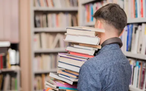 Mężczyzna w bibliotece niesie stos książek, jakby szukał streszczenia "Nad Niemnem".