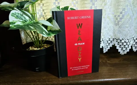 Książka "48 praw władzy" Roberta Greene'a na drewnianej półce, obok rośliny w czarnej doniczce.