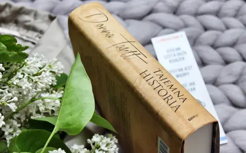 Książka "Tajemna historia" Donny Tartt na tle szarego, plecionego koca, obok gałązki bzu.