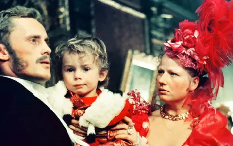 Rodzina w bogatych strojach, ojciec trzyma dziecko. Scena z filmu "Ziemia obiecana", streszczenie ukazuje losy bohaterów.