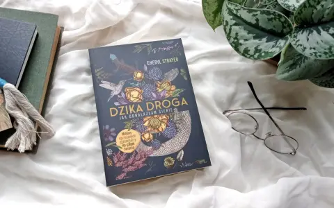 Okładka książki "Dzika droga" z motywami roślinnymi i zwierzęcymi. Obok leżą okulary i inne książki.