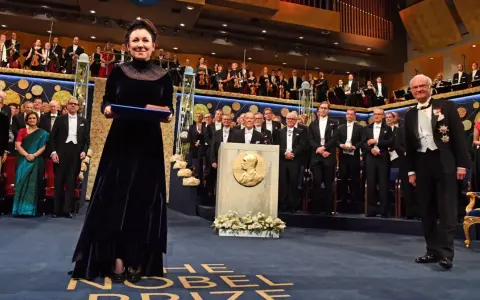 Uroczystość wręczenia Nagrody Nobla. W centrum kobieta w granatowej sukni z dyplomem. W tle orkiestra i laureaci. Tokarczuk nobel za co? Za literaturę.
