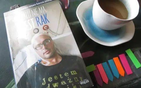 Książka "Żalu nie ma, jest rak" z wizerunkiem autora, obok filiżanka kawy i kolorowe zakładki.