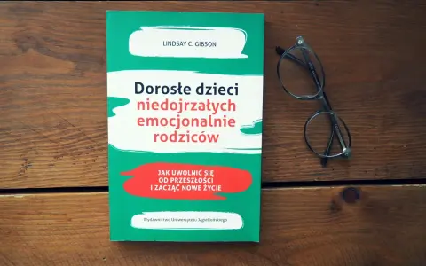 Książka "Dorosłe dzieci niedojrzałych emocjonalnie rodziców" i okulary na drewnianym tle.