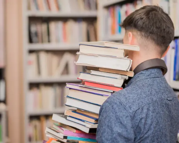 Mężczyzna w bibliotece niesie stos książek, jakby szukał streszczenia "Nad Niemnem".