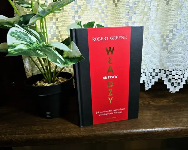 Książka "48 praw władzy" Roberta Greene'a na drewnianej półce, obok rośliny w czarnej doniczce.