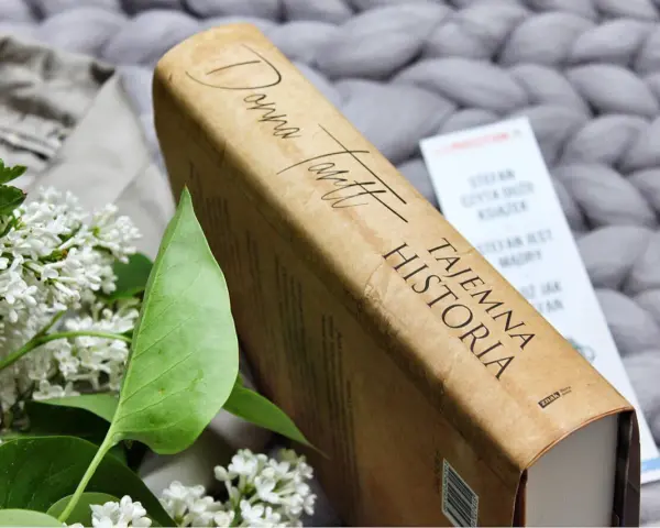 Książka "Tajemna historia" Donny Tartt na tle szarego, plecionego koca, obok gałązki bzu.
