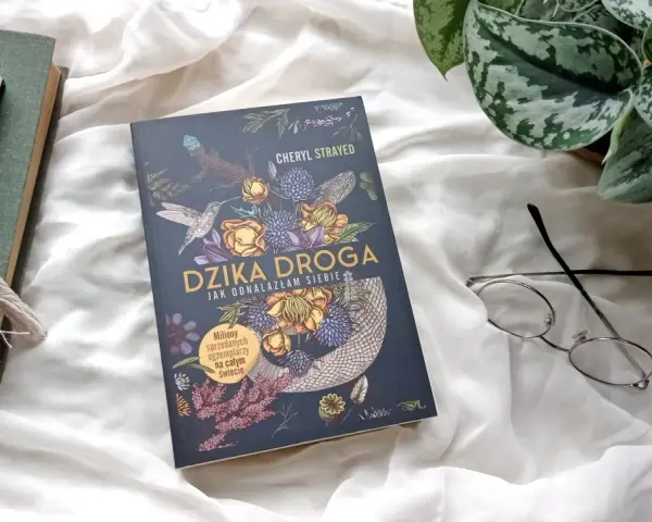 Okładka książki "Dzika droga" z motywami roślinnymi i zwierzęcymi. Obok leżą okulary i inne książki.