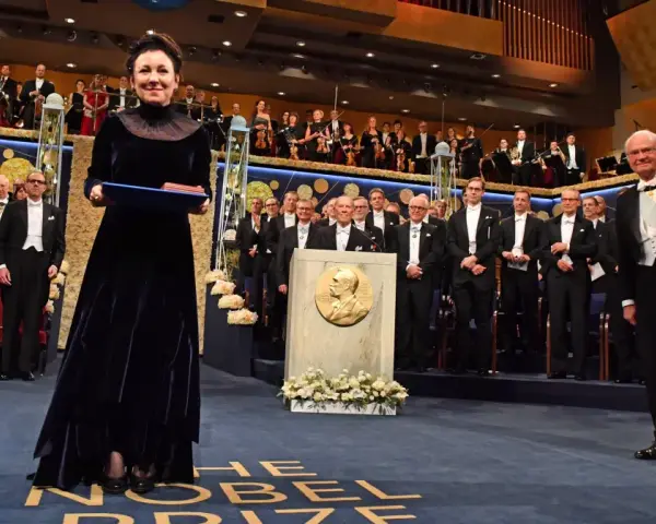 Uroczystość wręczenia Nagrody Nobla. W centrum kobieta w granatowej sukni z dyplomem. W tle orkiestra i laureaci. Tokarczuk nobel za co? Za literaturę.