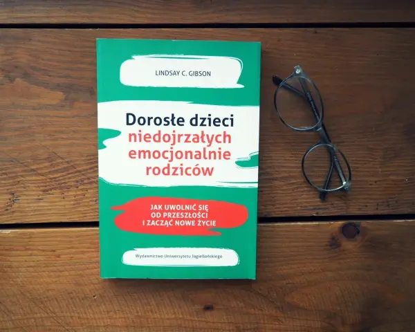 Książka "Dorosłe dzieci niedojrzałych emocjonalnie rodziców" i okulary na drewnianym tle.
