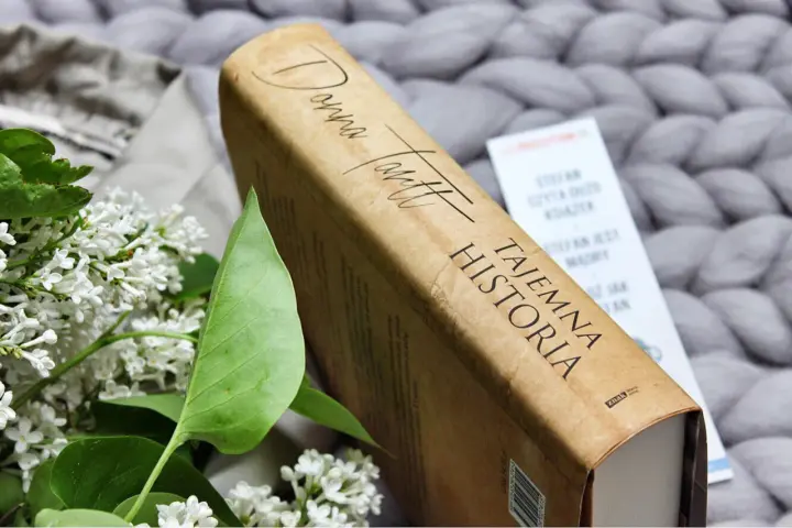 Książka "Tajemna historia" Donny Tartt na tle szarego, plecionego koca, obok gałązki bzu.