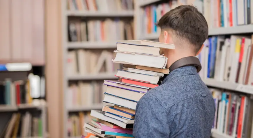 Mężczyzna w bibliotece niesie stos książek, jakby szukał streszczenia "Nad Niemnem".