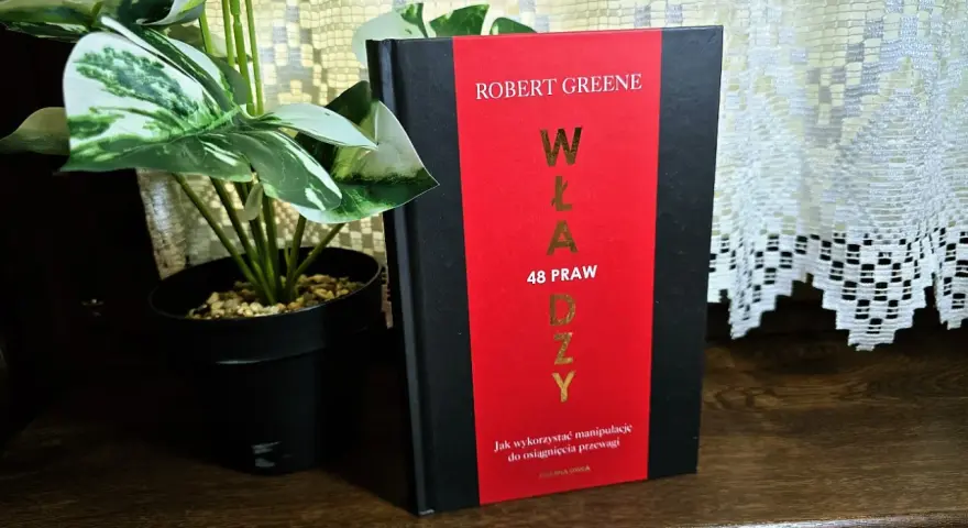 Książka "48 praw władzy" Roberta Greene'a na drewnianej półce, obok rośliny w czarnej doniczce.