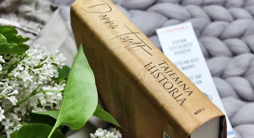 Książka "Tajemna historia" Donny Tartt na tle szarego, plecionego koca, obok gałązki bzu.