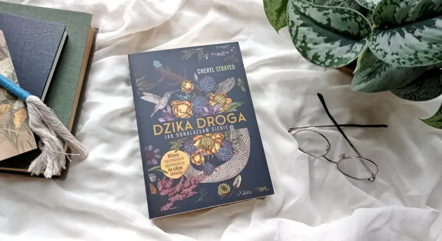 Okładka książki "Dzika droga" z motywami roślinnymi i zwierzęcymi. Obok leżą okulary i inne książki.