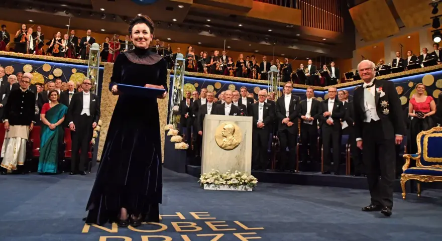 Uroczystość wręczenia Nagrody Nobla. W centrum kobieta w granatowej sukni z dyplomem. W tle orkiestra i laureaci. Tokarczuk nobel za co? Za literaturę.
