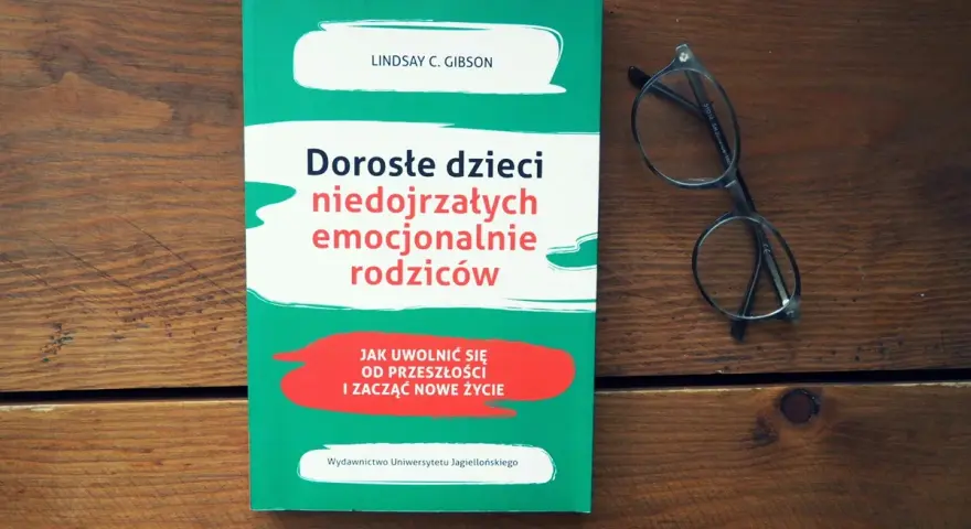 Książka "Dorosłe dzieci niedojrzałych emocjonalnie rodziców" i okulary na drewnianym tle.