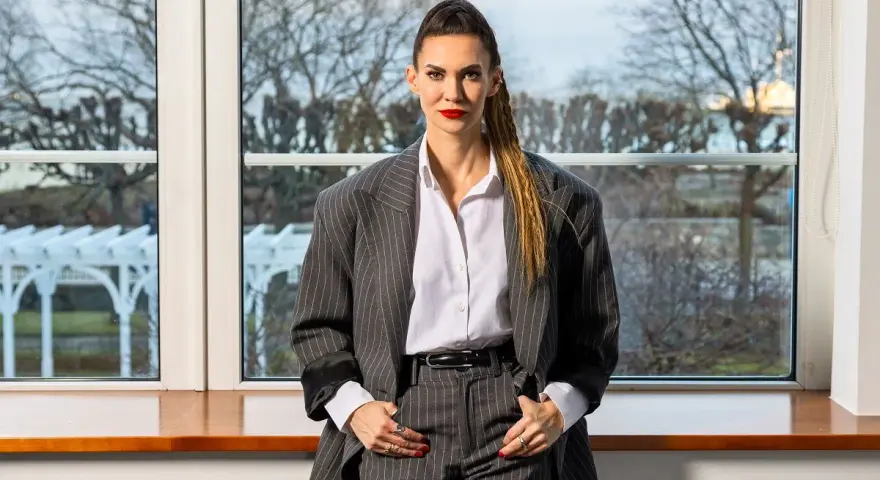 Małgorzata Oliwia Sobczak w garniturze, z włosami spiętymi w kucyk. Za nią okno z widokiem na drzewa, jakby szukała inspiracji do kolejnych książek.