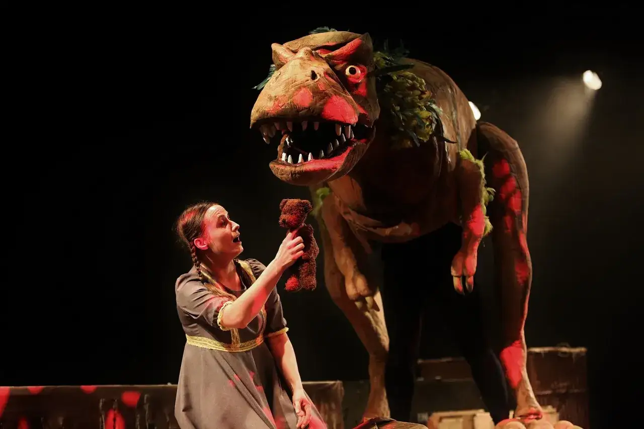 Dziewczynka z misiem staje naprzeciw groźnego dinozaura na scenie. To żywy obraz, jakby z teatrum mundi, gdzie każdy odgrywa swoją rolę.