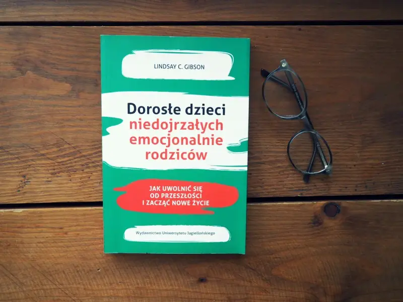 Książka "Dorosłe dzieci niedojrzałych emocjonalnie rodziców" i okulary na drewnianym tle.